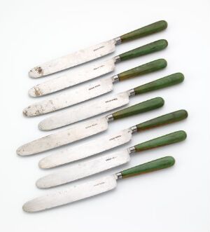  A set of seven vintage table knives with green handles and tarnished blades arranged in a slight fan shape on a white background.