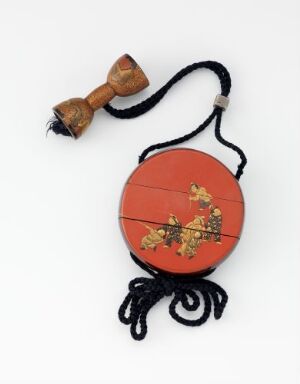  A traditional red inro with a golden floral design accompanied by a black braided cord with thick tassels and a small wooden netsuke. The background is off-white, emphasizing the object's color and design.