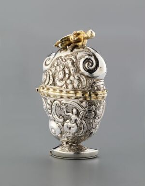  An intricately designed silver container with elaborate scrollwork and acanthus patterns, featuring a gold-colored figure or finial on top, set against a neutral gray background.