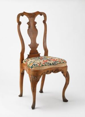  An antique wooden chair with an ornately carved back and cabriole legs. The chair has a thick, floral-patterned cushion in red, green, yellow, and beige on its seat.