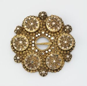  An intricately designed circular gold brooch with a central eyelet, surrounded by symmetrical patterns of faceted stones and smaller embellishments, creating a sparkling and ornate piece of jewelry.