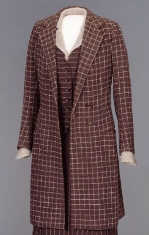  A vintage long coat and pleated skirt ensemble with a checkered pattern in dark and lighter brown, accented with off-white cuffs and collar, displayed against a neutral grey background.