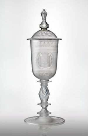  An elegant transparent glass vessel with a lid, featuring a detailed cylindrical container with an etched emblem, an artfully crafted stem with a series of bulbous and spindle-like sections, and a flat, round base, set against a gradient grey background. Artistname and title of the piece are unknown.