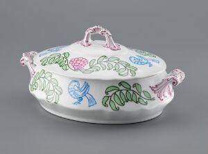  An oval-shaped feldspar porcelain covered serving dish designed by Oluf Wold-Torne, featuring a white background with a hand-painted overglaze decoration of colorful, stylized floral and faunal motifs on the lid, set against a neutral gray backdrop.