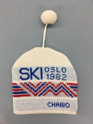  A white knitted hat with a white pom-pom, featuring blue and red knitting patterns and the text "SKI VM OSLO 1982" designed by Jan Søderstrøm.