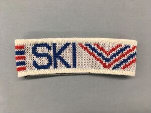  A knitted headband created by Jan Søderstrøm for the Ski World Championships in Oslo 1982, featuring the letters "SKI" in blue, two chevrons in blue and red, and the Roman numerals "VM" in red on an off-white background.