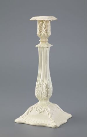  An ornate, pale-colored candlestick holder with a fluted column design and intricate leaf carvings on its base and top, standing against a neutral grey background.