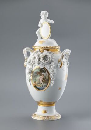  A finely detailed, white and gold porcelain vase with sculpted floral embellishments and a classical figure on top. The piece features a central medallion depicting a soft-colored pastoral scene.