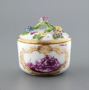  An ornate white porcelain covered box adorned with a hand-painted purple pastoral scene, gold gilt accents, and a three-dimensional floral bouquet on the lid, set against a neutral grey background.