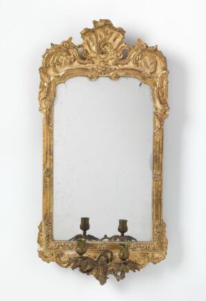  An antique, ornate, gold-framed mirror with a decorative crest on top and dual candle sconce at the bottom, set against a white background.