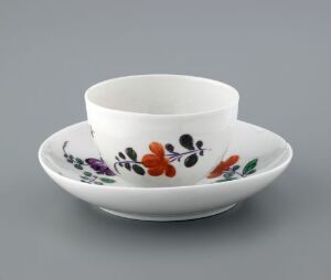  A white porcelain teacup without a handle placed in its matching saucer, both adorned with a delicate, hand-painted floral design featuring orange, purple, green, and hints of blue on a gray background.