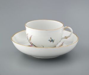  A porcelain teacup and matching saucer with a cream background, adorned with a thin gold rim, and a delicate pattern of soft purple and pink flowers, along with small blue and black insects, set against a light grey backdrop.