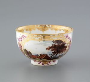  A porcelain bowl with a flared rim and a small base, featuring a brown pastoral landscape with touches of green around the exterior, accented by gold designs and colorful floral or symbolic motifs above, set against a grey background.