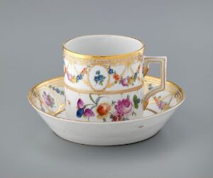  An elegant porcelain teacup and saucer set on a gray background. The set features vibrant, multicolored flower patterns with an overarching gold band and intricate golden embellishments on the rim. The handle of the cup is also gilded in gold, complementing the luxurious design.