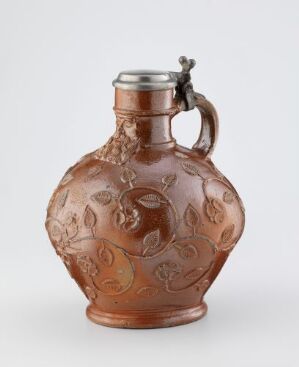  An amber-colored glass flask with intricate floral relief and a contrasting metal stopper with a mounted figure, featuring a curving handle that echoes the relief design.