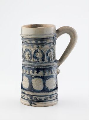  A cylindrical ceramic mug with an ornate blue and lighter blue decorative band featuring arch-like motifs and figures, framed by top and bottom bands in a natural, off-white ceramic color, with a sturdy handle attached on one side.