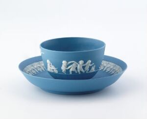  A mid-tone blue ceramic bowl with a white sculptural frieze of figures around the exterior, placed inside a matching blue plate with a similar frieze on its rim, set against a light gray background.