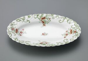  An oval porcelain dish with floral print design, featuring small clusters of flowers in shades of pink, red, green, and yellow, with a rim decorated with green swirl patterns on a glossy white background. Artist: Ukjent, Title: Unknown.