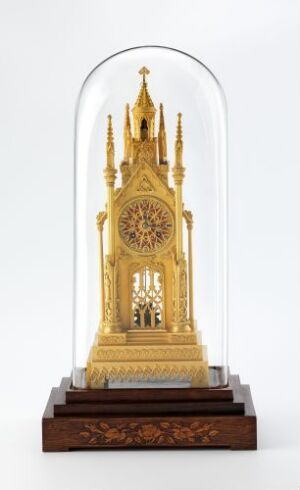  A miniature golden architectural structure with Gothic elements housed under a clear glass dome on a carved wooden base set against a white background. Artistname and title are unknown.