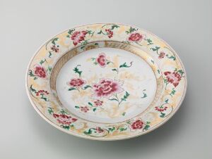  A decorative ceramic plate with a creamy off-white base, featuring a detailed floral pattern with a central cluster of pink roses surrounded by smaller flowers and foliage, and a richly decorated rim with deep pink and red flowers against a subtle peach-toned background. Artistname and title are unknown.