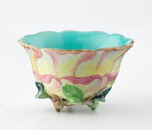  A decorative ceramic bowl with a fluted edge and a glazed turquoise interior. The exterior features soft yellow and pink stripes with green accents, supported by brown and green branch-like elements at the base.