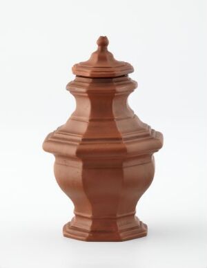  A terracotta-colored ceramic pot with a lid and decorative handle, displayed against a stark white background.