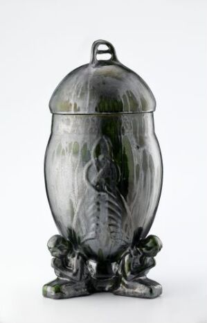  A decorative urn with a gray metallic sheen and a ribbed, vertical pattern on the body. It has a domed lid with a loop handle and is embellished with leaf-like elements at the base, against a neutral background. Artist name and title unknown.