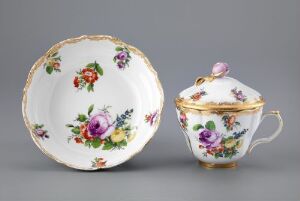  A porcelain tea set with a floral pattern on a gray background, consisting of a scalloped saucer and a teacup with a lid, both adorned with richly colored flowers and gold accents.