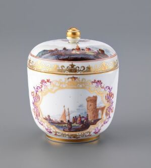  An oval-shaped porcelain box, with a painted maritime landscape scene in browns, blues, and greens wrapping around the center. It features gold scrollwork and magenta accents, and a gold knob on the lid.