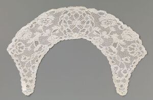  A crescent-shaped piece of white lace with floral patterns, displayed on a plain gray background. Artistname and title are unknown.