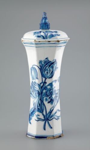  An antique hexagonal white ceramic jar with a lid, featuring vibrant blue floral patterns, displayed against a grey background.