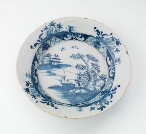  An antique round ceramic plate with a blue and white pastoral landscape scene within an oval frame, surrounded by elaborate floral patterns, set against a white background.