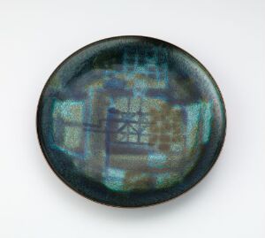  Circular iridescent object with shimmering blues, greens, purples, and teals, featuring a central emblem that appears distorted, possibly a geometric or floral design, against a plain background. Artist name and title are unknown.