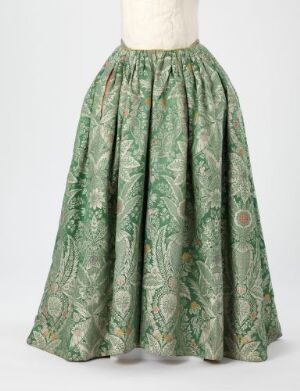  A voluminous, antique full-length skirt in sage green with detailed lighter green, gold, and red patterns, displayed on an off-white mannequin against a light background.