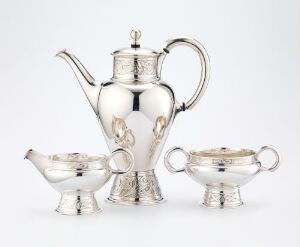  A set of polished silver teaware on a white background, consisting of a teapot with a curved spout and handle, a creamer, and a lidded sugar bowl, all gleaming with reflected light.