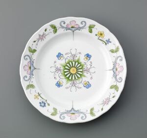  A decorative ceramic plate with scalloped edges, featuring a pastel-hued floral pattern on the rim, and a symmetrical, radiating design in green, yellow, and blue at the center.