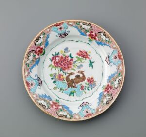  An ornate decorative plate with a pale blue background, featuring a central image of a bird perched on a branch surrounded by vibrant pink, red, and white flowers, and bordered by gold-rimmed floral accents and small butterflies.