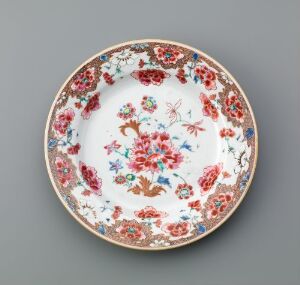  Decorative porcelain plate featuring intricate floral designs in red, blue, green, and yellow hues with a gold or peachy rim, against a neutral gray background.