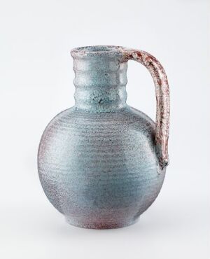  "Mugge" by Louis Benjamin, a glazed earthenware mug with a bulbous body and tapered neck in shades of blue-grey to teal, featuring a contrasting reddish-brown handle. The image shows the textured surface of the ceramic with a white background.