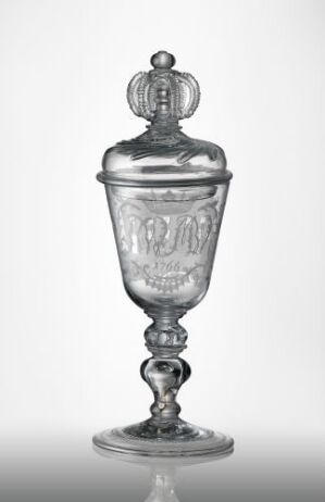  An ornate clear glass goblet with a lid set against a light gray background. The goblet features a rounded finial, an etched bowl, a decorative stem, and a flared base, displaying the intricate detailing typical of elegant antique glassware.