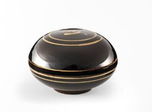  A polished black spherical container with a lid, adorned with three thin horizontal golden bands, set against a white background. Artist name and title are unknown.