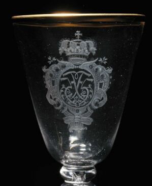 
 An ornate clear glass goblet with gold rim, featuring an intricate etched crest and monogram design, set against a black background.