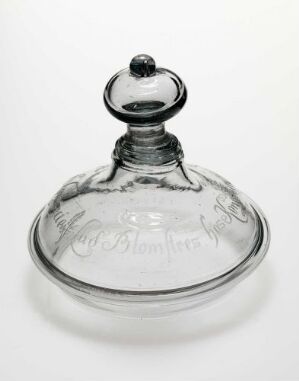 
 A clear glass butter dish with an etched inscription on the side of the base and a domed lid with a spherical knob on top, set against a white background. Artist name and title are unknown.