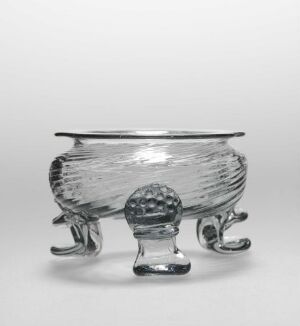  A small transparent glass footed bowl with a textured bottom half and smooth rim, supported by three curved feet, with a decorative knob in the center bottom, against a neutral gray background. Artist name and title remain unknown.