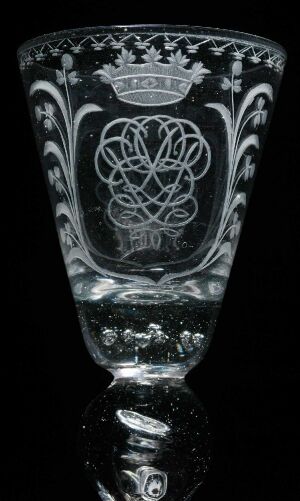  An intricately engraved transparent glass titled "Perlkelchen" by Nøstetangen glassverk, featuring a central heraldic emblem surrounded by floral and fleur-de-lis patterns, with a pearl-like detail along the upper rim. The piece exudes elegance through its monochromatic, light-reflective engravings.