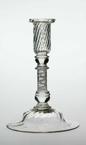  A clear glass candlestick with a broad base, decorative stem with spherical elements, and a ribbed candle cup, set against a light greyish background.