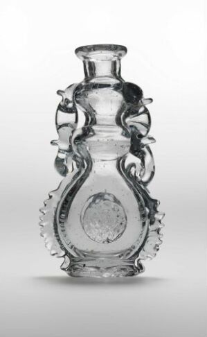  A transparent glass bottle or vase with intricate design features, including twisted handles, a sunburst pattern surrounding a central medallion, and a beaded texture on the lower part, set against a plain white background.