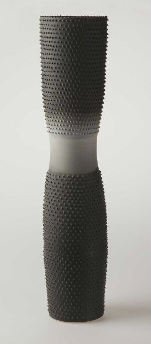  A cylindrical porcelain object titled "Krukke med pels" by artist Sidsel Hanum, with a textured surface resembling a soft fur, in shades of gray transitioning from light at the top to dark at the bottom, against a neutral background.