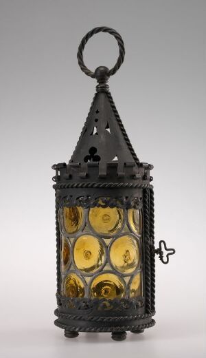  Antique-style metal lantern with intricate cut-out patterns and yellow glass panels, featuring a decorative ring at the top and an ornamental cross-shaped latch on the side, set against a neutral gray background.