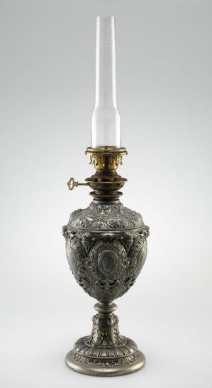  An elegantly detailed silver oil lamp with a brass burner and a tall white glass chimney, exhibiting intricate embossed patterns, floral motifs, and a classical design on a nondescript background.